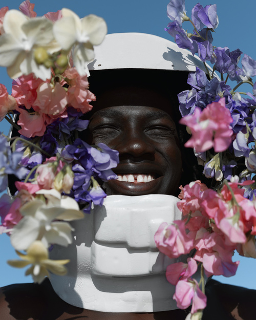 Close-up portrait of smiling adult with dark skin wearing a white helmet, surrounded by pink, purple, and white flowers against a blue sky.