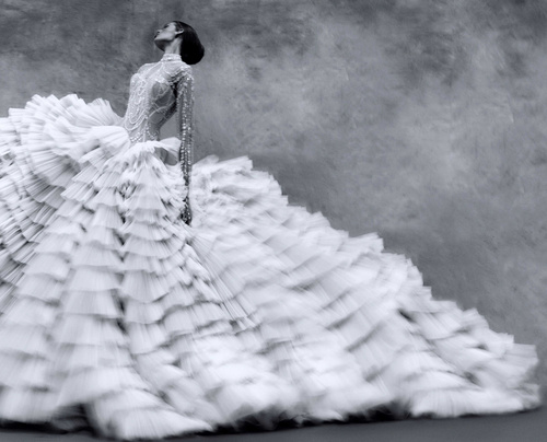 Woman posing in an elaborate white gown with ruffles in a studio with a gray backdrop, fashion editorial. Long exposure effect.