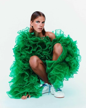 Model crouching wearing a voluminous green ruffled organza top and white sneakers against a plain white background.