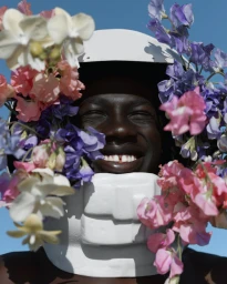 Close-up portrait of a Black man wearing a white helmet framed by pink, white, and purple flowers against a clear blue sky background.