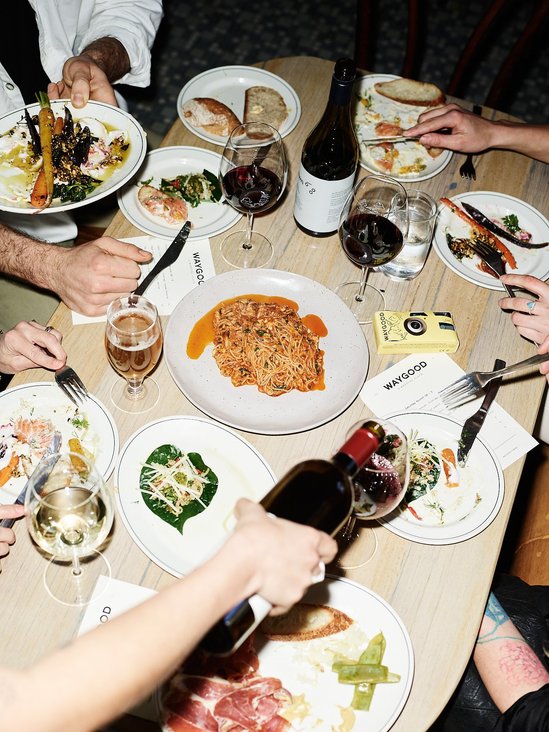 Overhead shot of a group of people dining, featuring plates of food, glasses of wine, and a yellow camera on a wooden table.