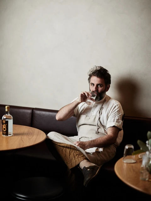 Man in an apron sits at a table, drinking from a glass in a warmly lit restaurant, with a bottle of gin and round tables visible.