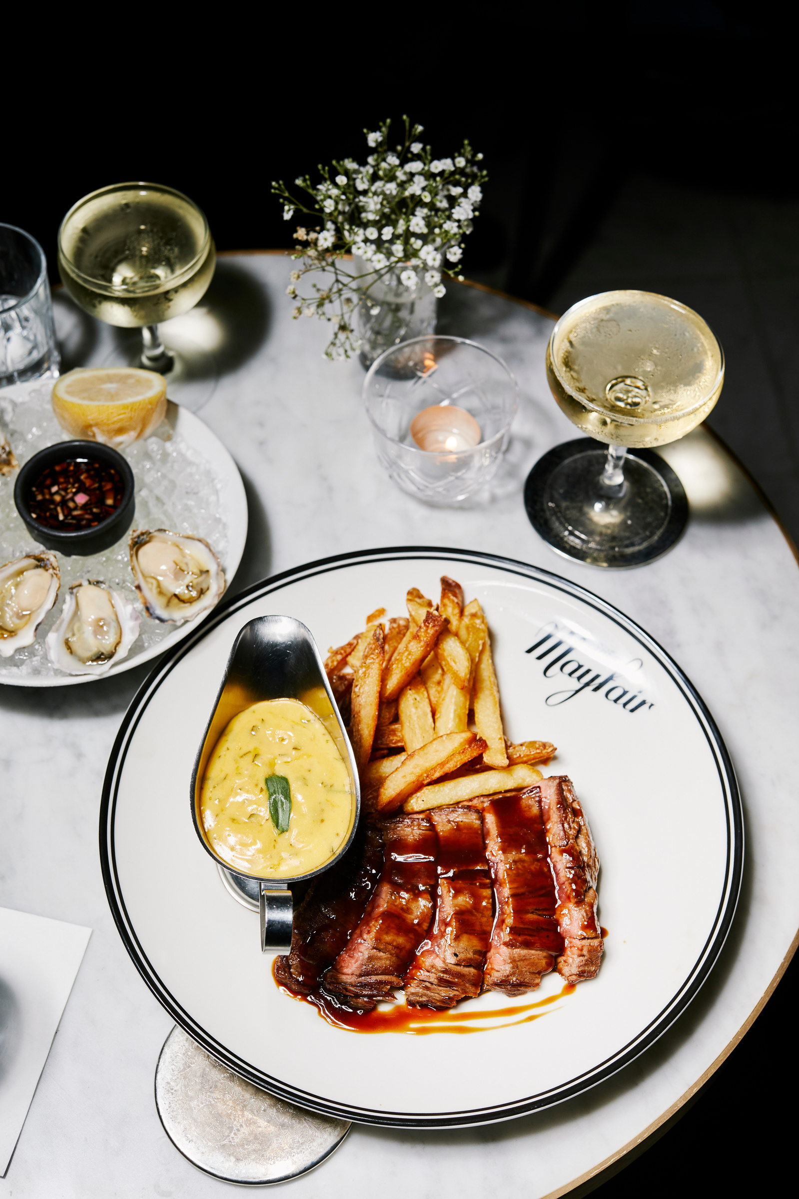 Overhead shot: A plate of sliced steak with fries and sauce, set on a marble table with champagne and oysters, inviting food photography.