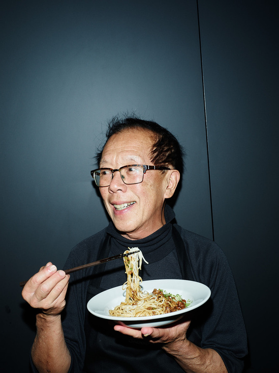 Smiling Asian man wearing glasses and a black apron holds a plate of noodles with chopsticks against a dark grey background indoors.