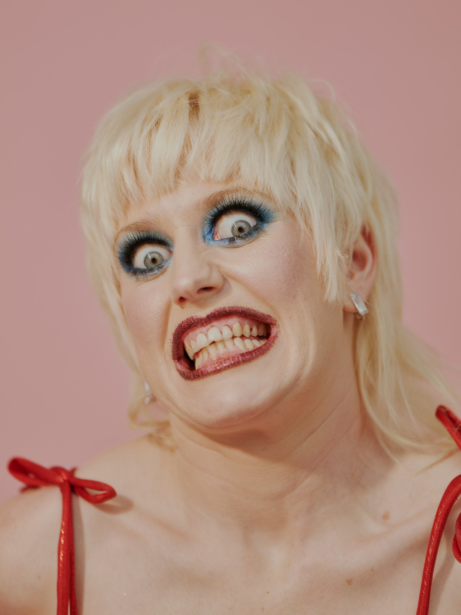 Close-up studio portrait of a white adult female with blonde hair and blue eye makeup, making a face with visible teeth against a pink background.