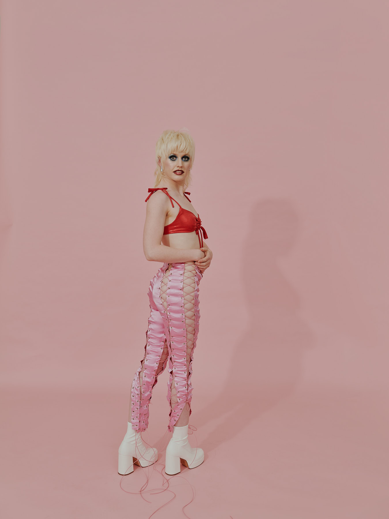 Full-length shot of a slender adult wearing a red bra top, laced pink pants and white platform boots in a pink photo studio.