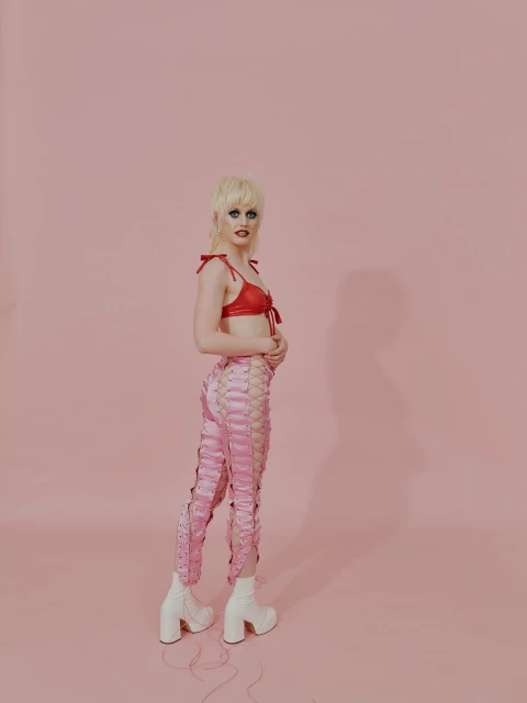 Full-length shot of a slender adult wearing a red bra top, laced pink pants and white platform boots in a pink photo studio.