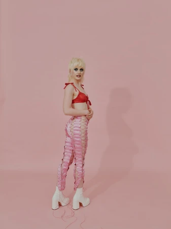 Full-length shot of a slender adult wearing a red bra top, laced pink pants and white platform boots in a pink photo studio.