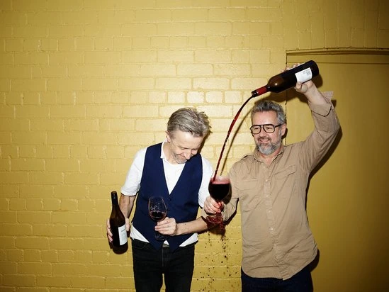Two adult men indoors are pouring red wine from a bottle into a glass, with spillage on the floor and a yellow brick wall background.