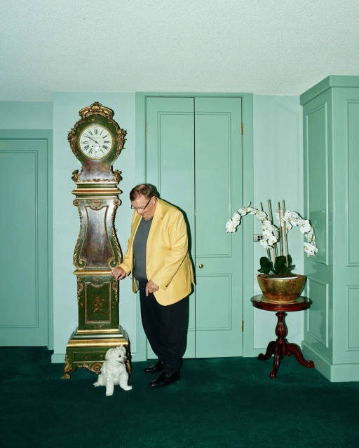 Man in a yellow jacket poses indoors with a white dog and a grandfather clock in a room with green walls and carpet.