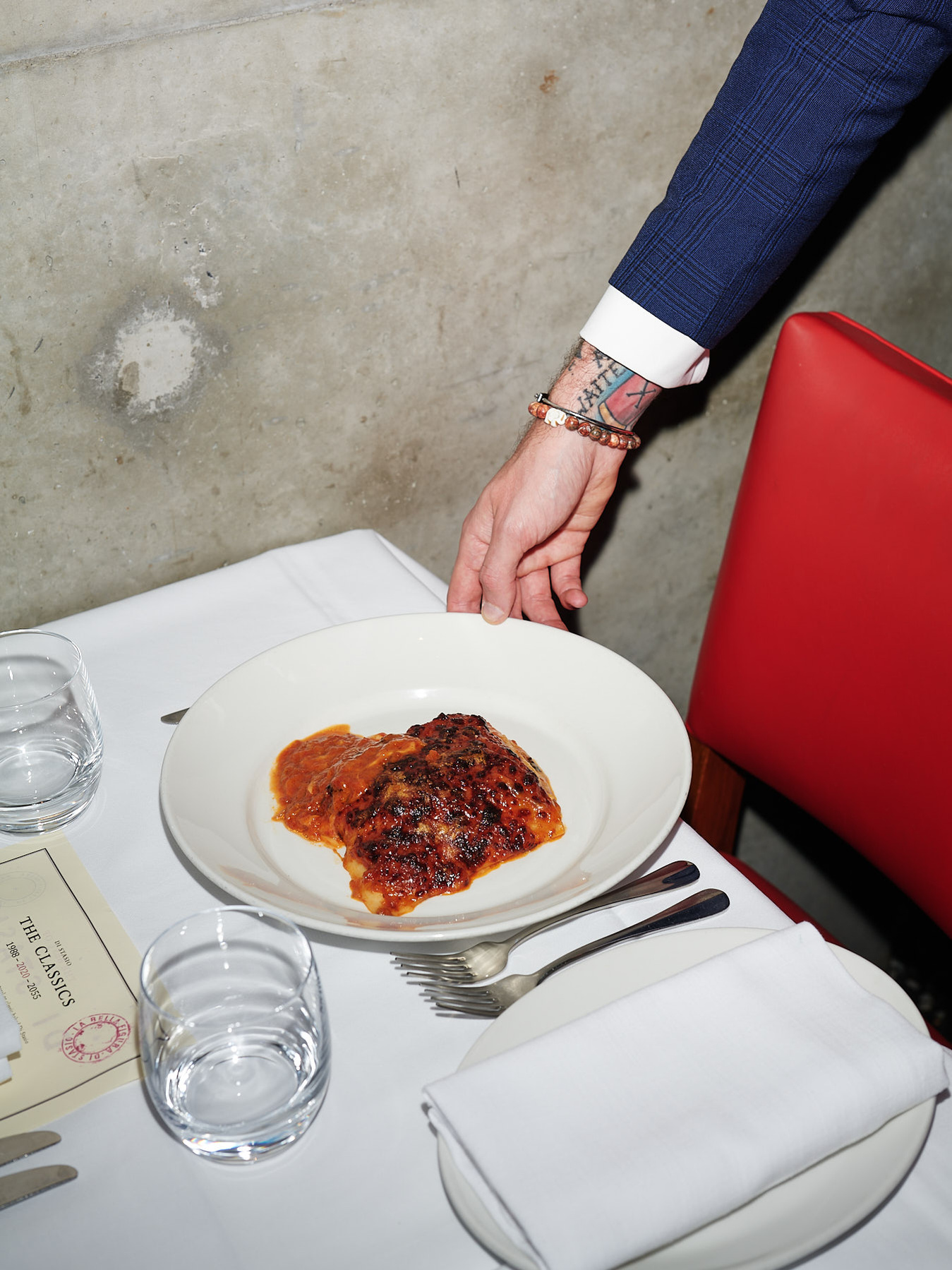 Overhead shot of plated lasagna on a restaurant table with glasses, silverware, and a blue-suited arm reaching for the plate.