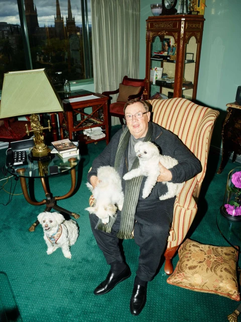 Man in eyeglasses sits in a striped chair in a living room, holding two small white dogs with two more white dogs on the green carpet.