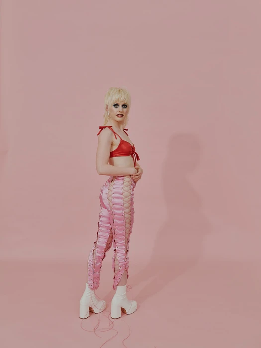 Model with blonde hair wearing a red top and pink lace-up pants against a solid pink backdrop