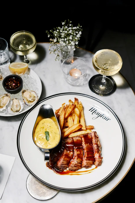 Overhead food photography of a steak frites dish with gravy boat, oysters, and two glasses of champagne on a marble table.