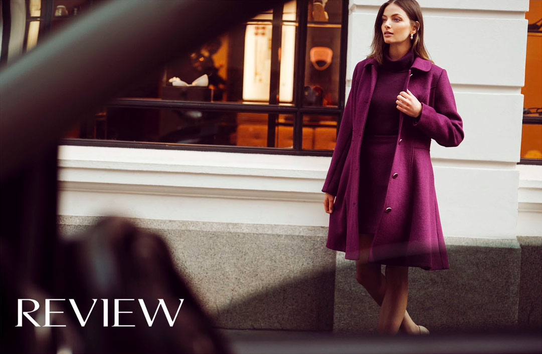 Woman in purple coat and dress stands outdoors against a white wall with brown window frames on a cloudy day.