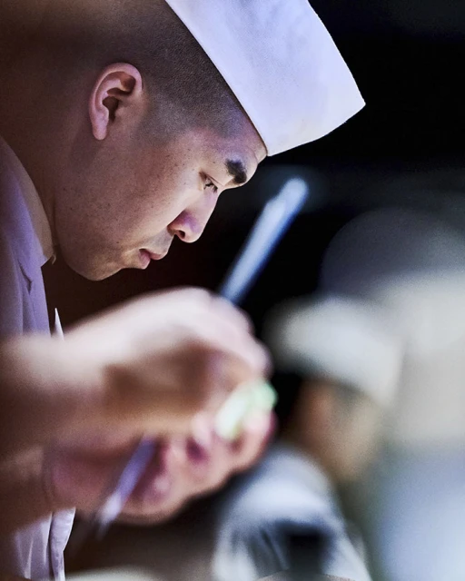 Profile of focused chef in white hat carefully prepping ingredients with chopsticks, set against a dark blurred background indoors.