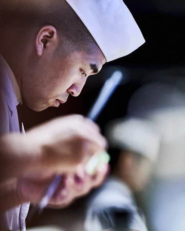 Profile of focused chef in white hat carefully prepping ingredients with chopsticks, set against a dark blurred background indoors.