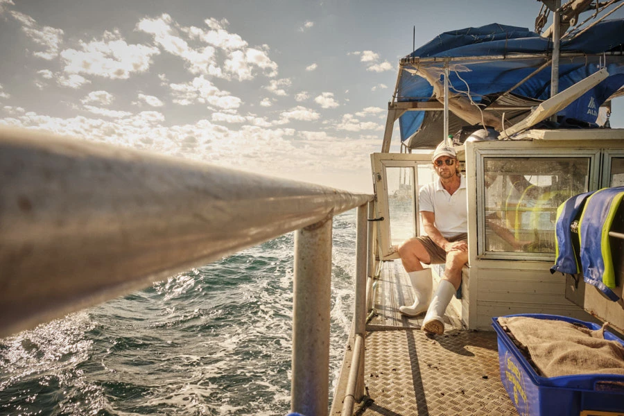 Adult man on fishing boat sits near blue container, ocean waves, and cloudy sky creating a bright, marine work environment.