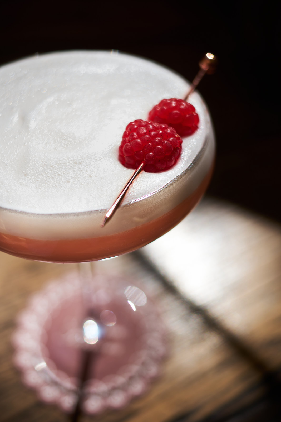 Elevated, close-up cocktail shot features a creamy white foam topped with two raspberries, served in a pink-stemmed glass.