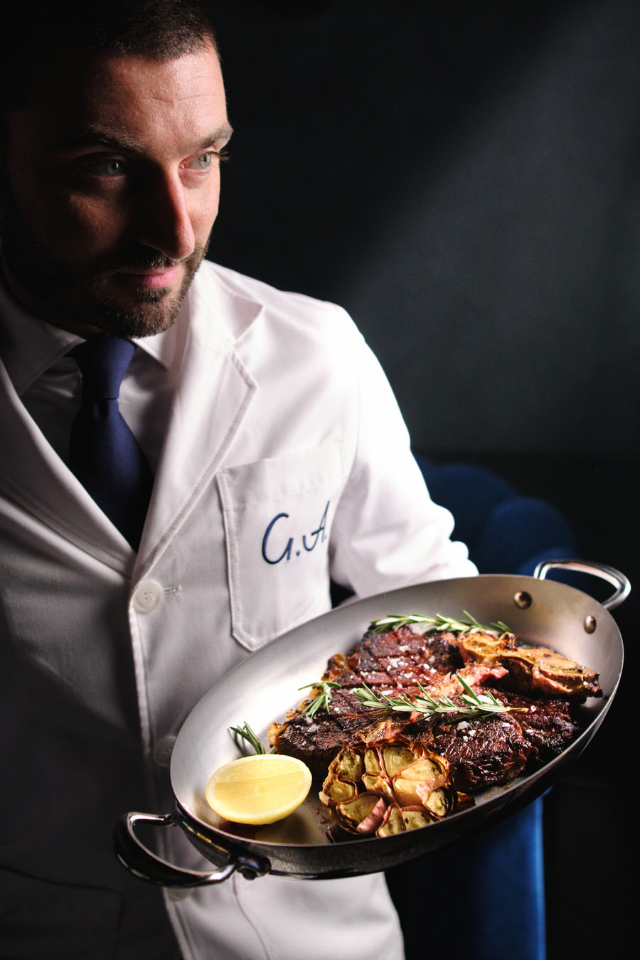 Adult male chef in a white coat holds a metal serving dish with cooked meat, potatoes, lemon, and rosemary sprigs on a black background.