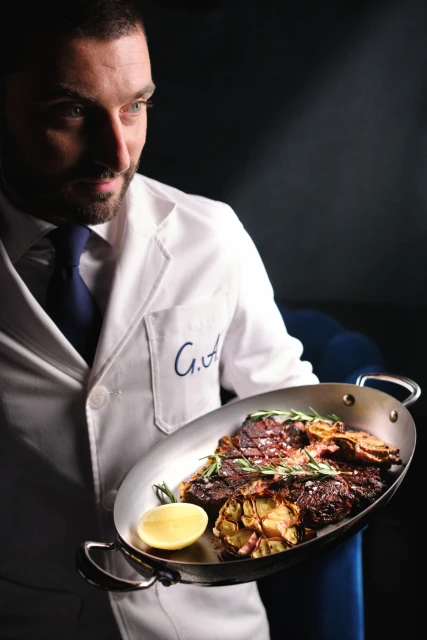 Adult male chef in a white coat holds a metal serving dish with cooked meat, potatoes, lemon, and rosemary sprigs on a black background.