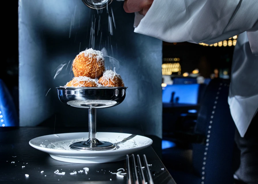Close-up of fried arancini balls on a silver stand being topped with grated cheese by a waiter in a dark restaurant setting.