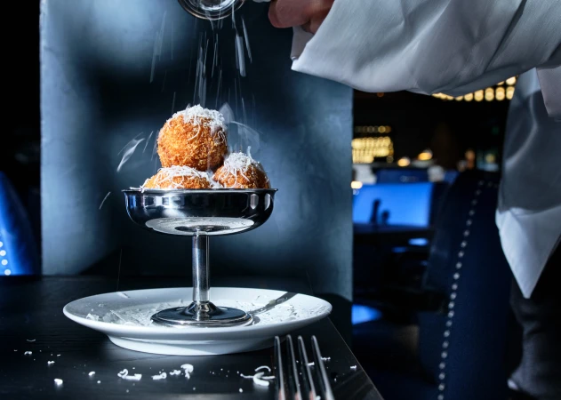Close-up of fried arancini balls on a silver stand being topped with grated cheese by a waiter in a dark restaurant setting.