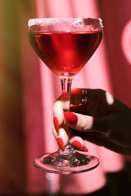 Close-up of a hand holding a red cocktail in a sugar-rimmed glass against a pink background with light and shadow patterns.