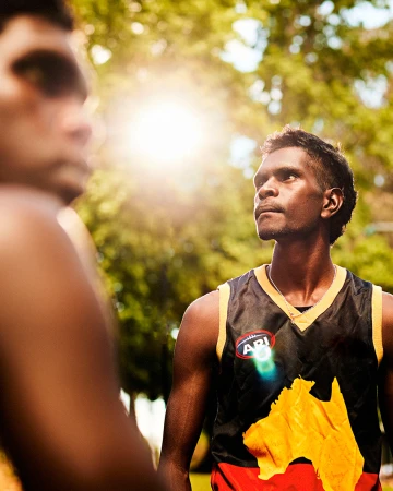Two Aboriginal Australian men outdoors in bright sunlight. One man in focus wearing a sports jersey, the other man out of focus.