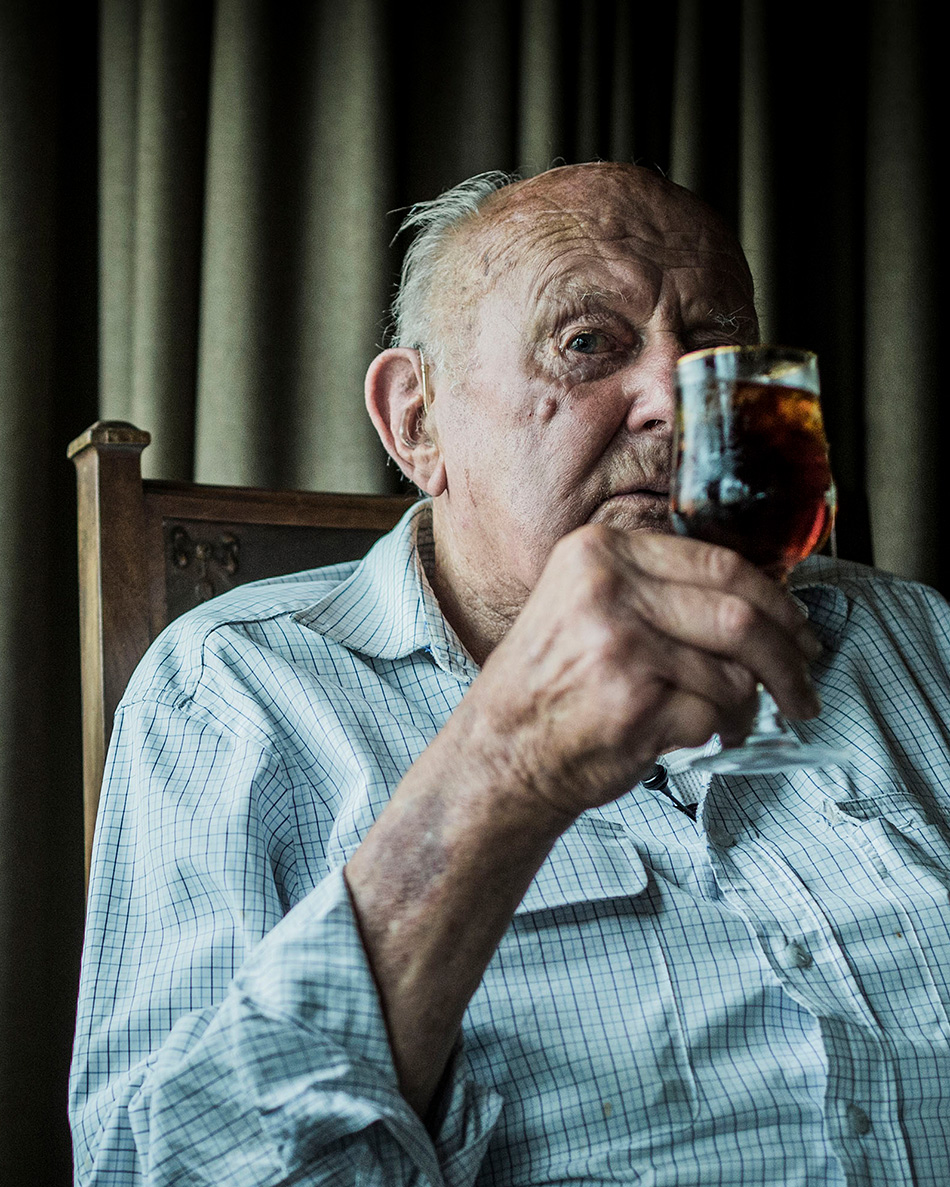 Elderly man in plaid shirt sips from a stemmed glass indoors, gazing directly at the camera with a serious expression. 
