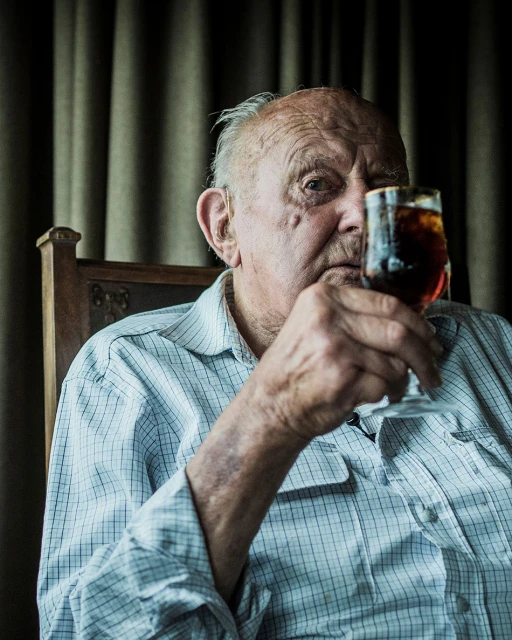 Elderly man in plaid shirt sips from a stemmed glass indoors, gazing directly at the camera with a serious expression. 