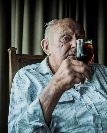 Elderly man in plaid shirt sips from a stemmed glass indoors, gazing directly at the camera with a serious expression. 