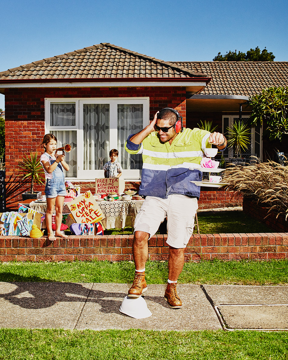 Adult male balancing tea set on tray while standing on hard hat; two children hosting tea party/fundraiser in front of brick house.