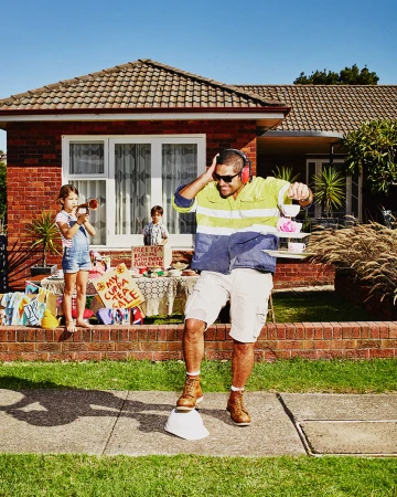Adult male balancing tea set on tray while standing on hard hat; two children hosting tea party/fundraiser in front of brick house.
