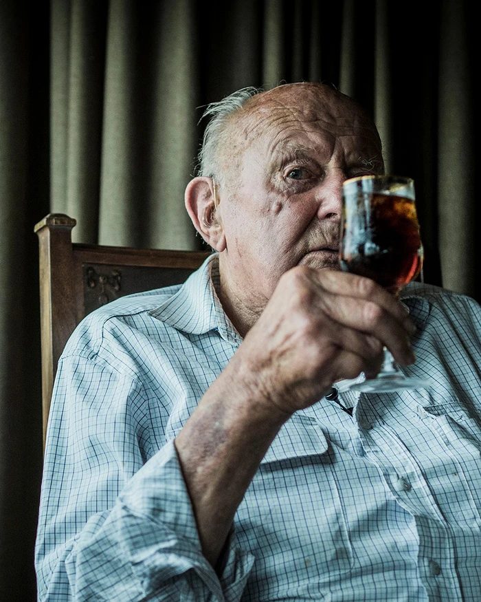 Elderly man in a plaid shirt seated indoors, holding a glass of dark beverage in front of his face.