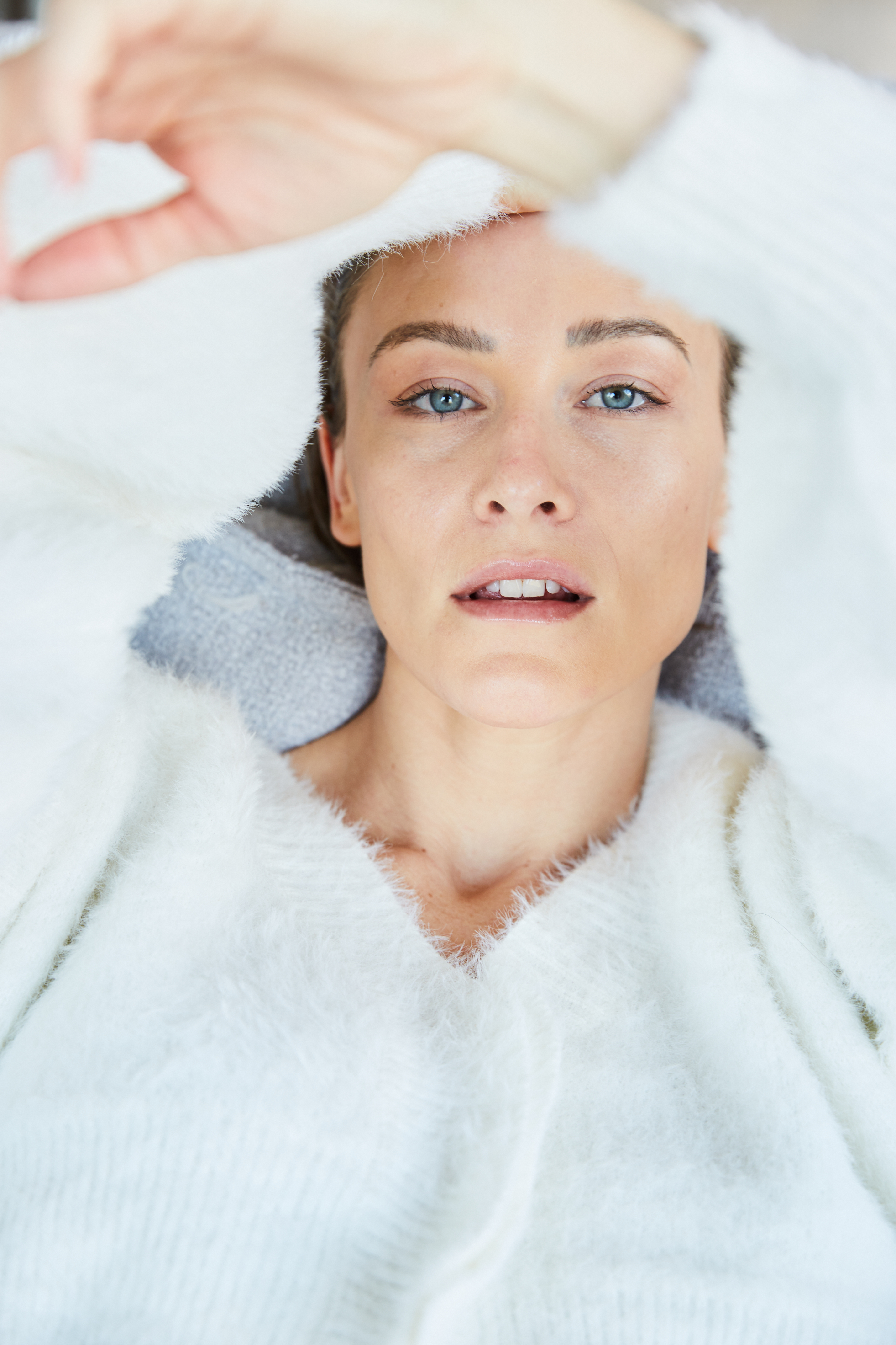 Close-up shot of an adult female wearing a fluffy white sweater looking at the camera with her hand raised above her head.