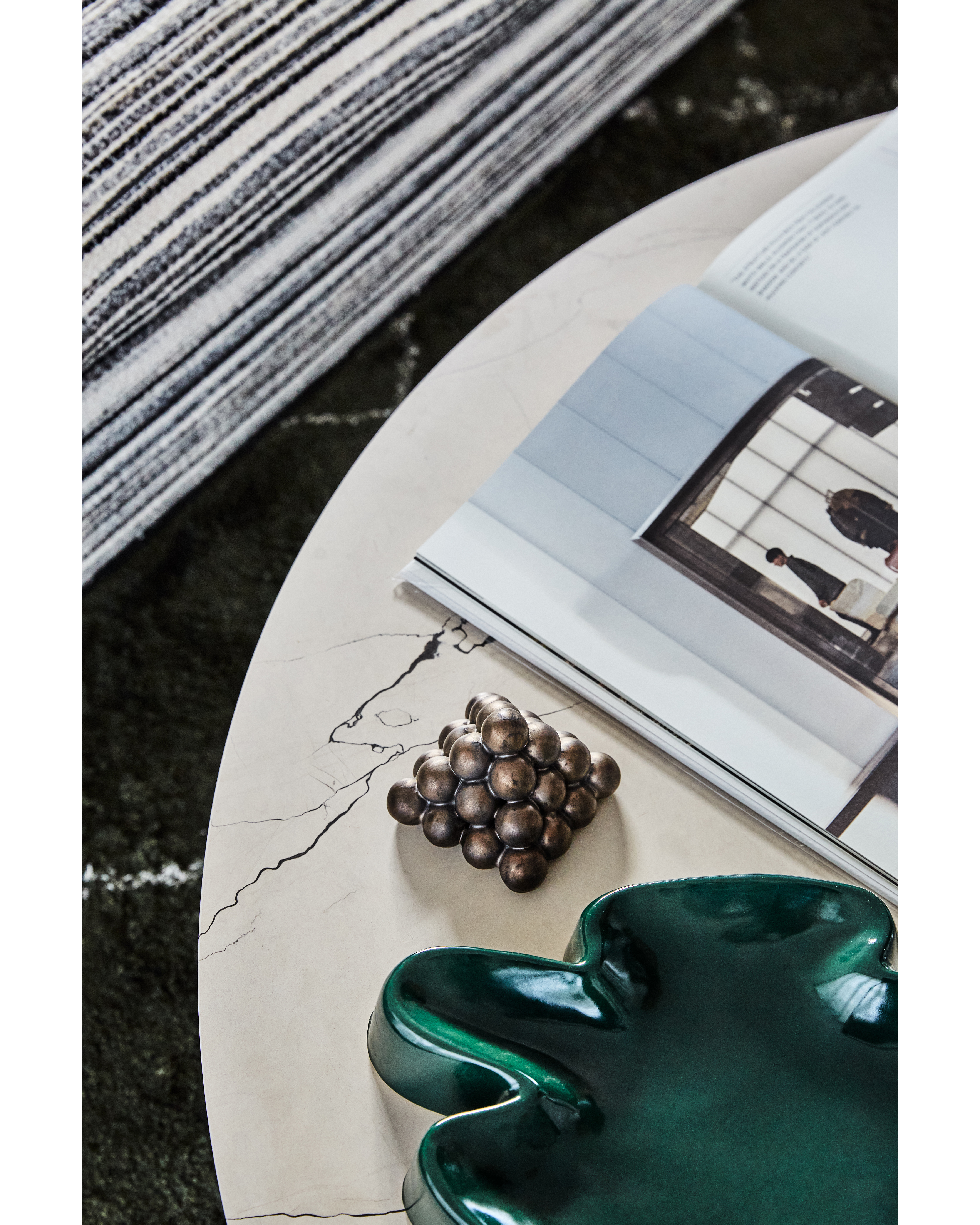 Close-up of a marble table displaying a magazine, decorative balls, and a green dish, set against a striped rug in an indoor setting.