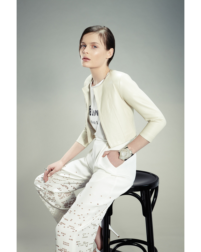 Fashion shot of a young female model in a light jacket and patterned pants, seated on a black stool against a neutral background.
