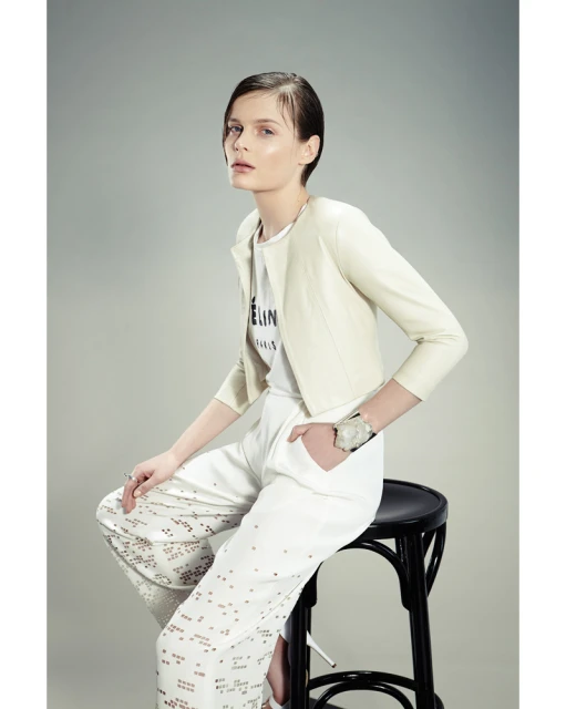 Fashion shot of a young female model in a light jacket and patterned pants, seated on a black stool against a neutral background.