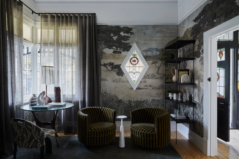Living room interior with wallpaper featuring nature scene, two olive striped chairs, round glass table, and diamond-shaped stained-glass window.