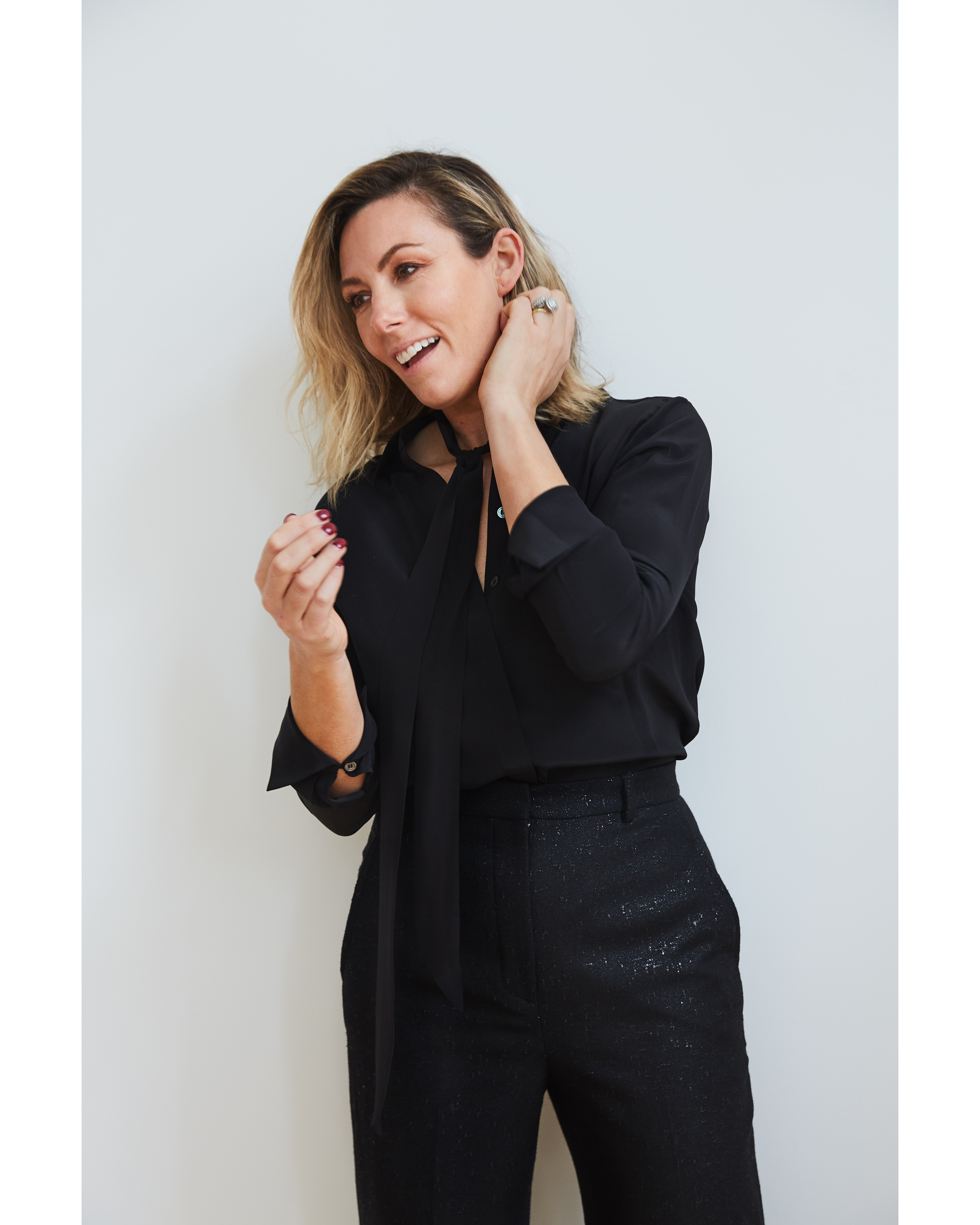 Blonde woman in black shirt and pants smiles and poses indoors against a white wall in a medium shot with an eye-level angle.