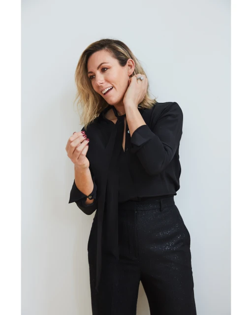 Blonde woman in black shirt and pants smiles and poses indoors against a white wall in a medium shot with an eye-level angle.