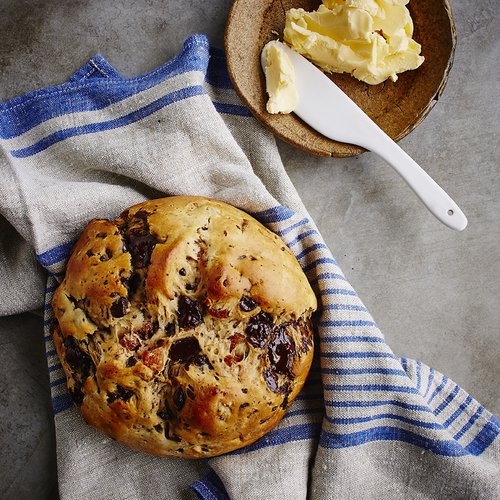 Overhead shot of a loaf of bread with dried fruit and herbs resting on a blue striped dish towel, served with butter and a knife.