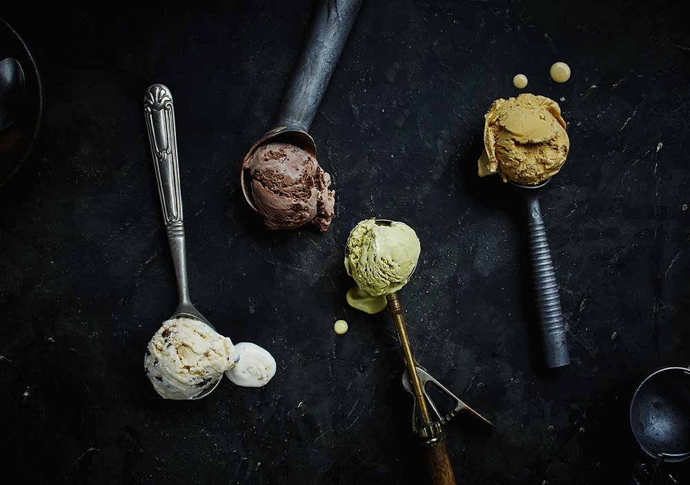Overhead shot of assorted ice cream scoops in metal serving spoons on a dark, textured surface, showcasing different flavors and textures.