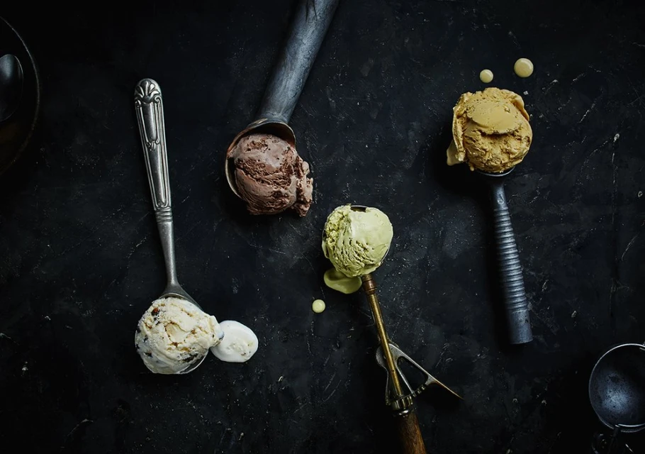 Overhead shot of assorted ice cream scoops in metal serving spoons on a dark, textured surface, showcasing different flavors and textures.