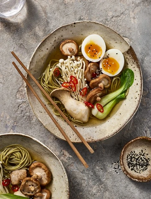 Overhead shot of two bowls of ramen with mushrooms, eggs, noodles, and greens on a textured gray surface. Japanese food.