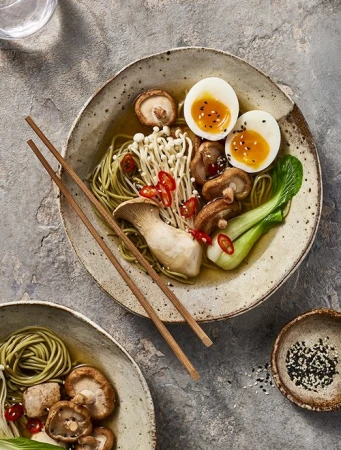 Overhead shot of two bowls of ramen with mushrooms, eggs, noodles, and greens on a textured gray surface. Japanese food.