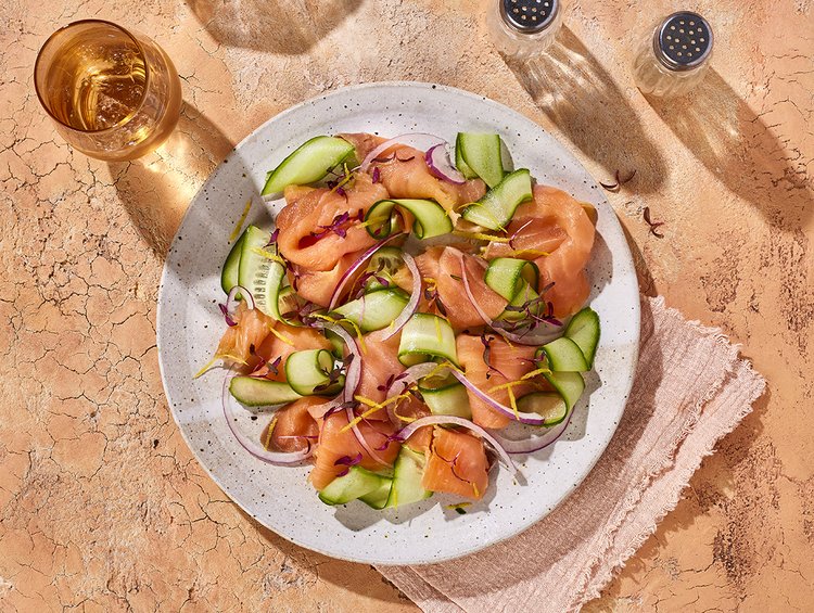 Overhead shot of a plate of salmon salad with cucumber and red onion, accompanied by a glass of liquid and salt and pepper shakers.