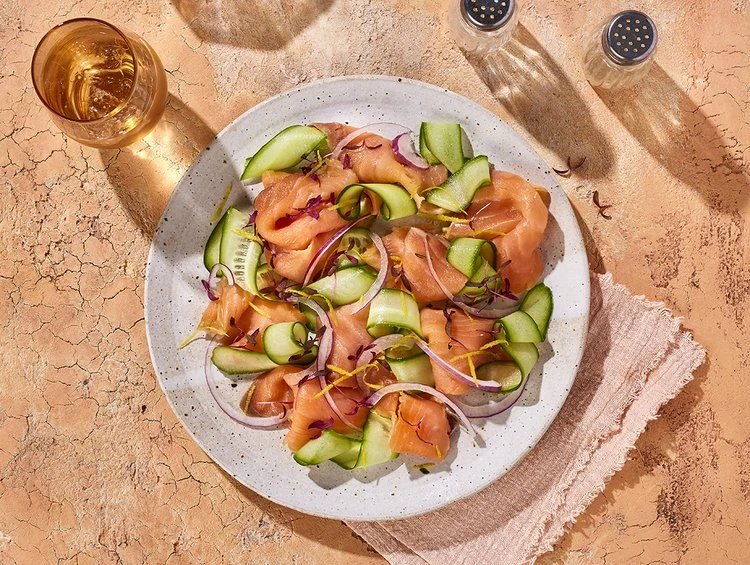 Overhead shot of a plate of salmon salad with cucumber and red onion, accompanied by a glass of liquid and salt and pepper shakers.