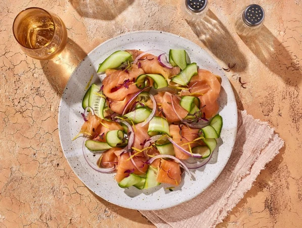 Overhead shot of a plate of salmon salad with cucumber and red onion, accompanied by a glass of liquid and salt and pepper shakers.